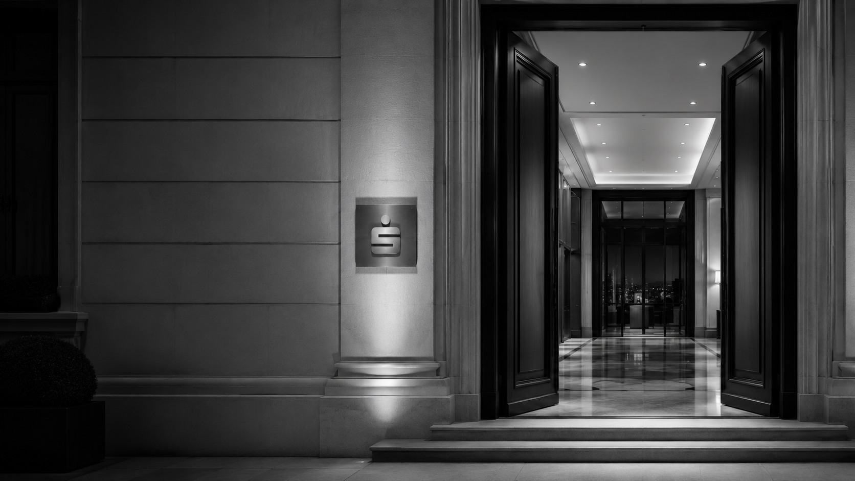 Beleuchteter Eingang einer Sparkasse bei Nacht mit dem S-Signet an der Steinfassade